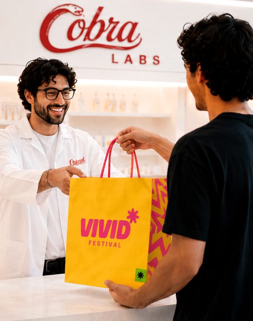 Operator in a white Cobra coat handing a Microsoft Ignite shopping bag to a guest at the pickup counter, framed by the backlit Cobra Labs wordmark.